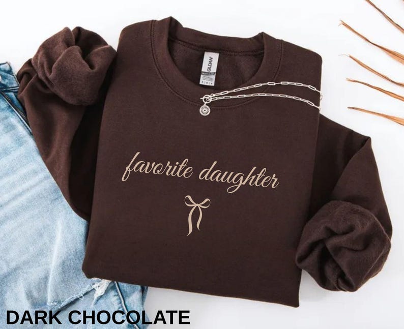 Puede incluir: Una sudadera de color chocolate oscuro con las palabras "favorite daughter" en una fuente cursiva, junto con un gr&aacute;fico de lazo. Un collar plateado con un colgante est&aacute; drapeado en la parte delantera. El texto "DARK CHOCOLATE" est&aacute; en la parte inferior.