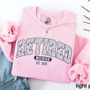 May include: A light pink sweatshirt with the word "RETIRED" in a navy blue, floral-patterned font. Below the word "RETIRED" is the word "NURSE" and the year "EST. 2026". The sweatshirt is folded on top of a pair of blue jeans.