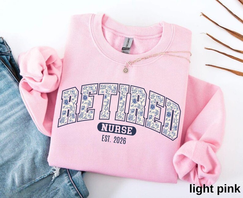 May include: A light pink sweatshirt with the word "RETIRED" in a navy blue, floral-patterned font. Below the word "RETIRED" is the word "NURSE" and the year "EST. 2026". The sweatshirt is folded on top of a pair of blue jeans.
