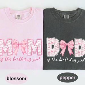 May include: A light pink t-shirt with the text MOM of the birthday girl and a dark charcoal t-shirt with the text DAD of the birthday girl. Both shirts feature a large pink bow graphic replacing the letter O in MOM and the letter A in DAD.