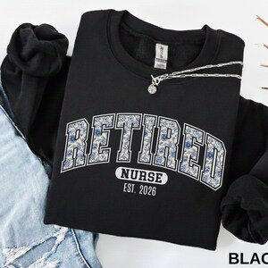 May include: Black sweatshirt with the word "RETIRED" in a blue floral design, arched across the chest. Below the word "RETIRED" is the text "NURSE EST. 2026". A silver necklace with a small pendant is draped across the sweatshirt.