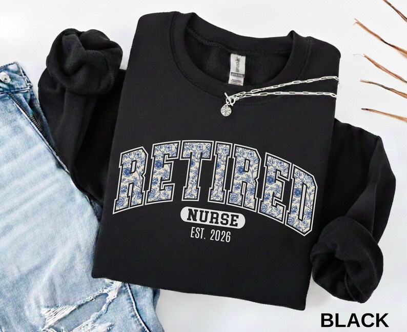 May include: Black sweatshirt with the word "RETIRED" in a blue floral design, arched across the chest. Below the word "RETIRED" is the text "NURSE EST. 2026". A silver necklace with a small pendant is draped across the sweatshirt.