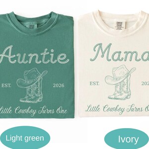 May include: A light green t-shirt and an ivory t-shirt, each featuring a graphic of a cowboy hat and boots. The green shirt has white text reading Auntie, EST. 2026, and Little Cowboy Turns One. The ivory shirt has green text reading Mama, EST. 2026, and Little Cowboy Turns One.
