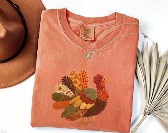 May include: A coral-colored t-shirt featuring a turkey applique with patchwork feathers in shades of brown, orange, and green. The shirt has a round neckline and a tag that reads "Comfort Colors". A gold necklace with a pendant is draped across the shirt.