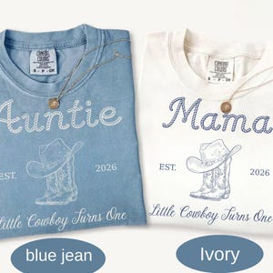 May include: A blue jean and an ivory t-shirt, each featuring a graphic of a cowboy hat and boots. The blue shirt displays the text Auntie, while the ivory shirt displays Mama. Both shirts include the text EST. 2026 and Little Cowboy Turns One.
