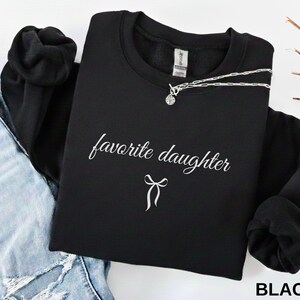 Puede incluir: Una sudadera negra con la frase "favorite daughter" en escritura blanca y un lazo. Un collar plateado con un colgante est&aacute; colocado sobre la sudadera. La palabra "BLACK" est&aacute; impresa en la esquina inferior derecha.