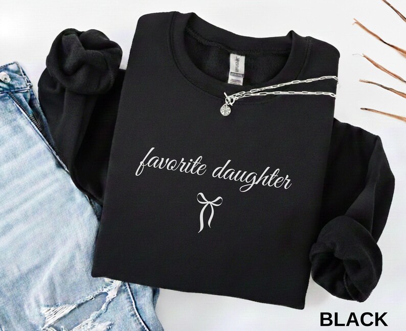 Puede incluir: Una sudadera negra con la frase "favorite daughter" en escritura blanca y un lazo. Un collar plateado con un colgante est&aacute; colocado sobre la sudadera. La palabra "BLACK" est&aacute; impresa en la esquina inferior derecha.