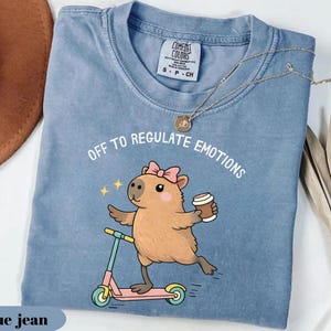May include: Blue jean Comfort Colors t-shirt featuring a cartoon capybara on a pink scooter, holding a coffee cup. The text "OFF TO REGULATE EMOTIONS" is above the capybara. A gold necklace is draped across the shirt.