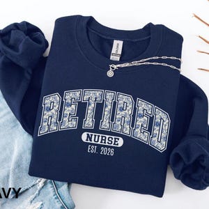 May include: Navy blue sweatshirt with the word "RETIRED" in large white block letters with a floral pattern. Below the word "RETIRED" is the word "NURSE" and the year "EST. 2026". A silver necklace with a pendant is draped across the sweatshirt.