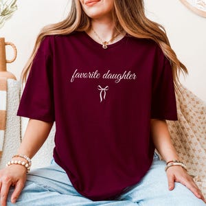 Puede incluir: Camiseta granate con la frase "favorite daughter" en cursiva y un peque&ntilde;o lazo. La camiseta es de color liso y tiene mangas cortas.