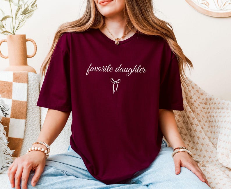 Puede incluir: Camiseta granate con la frase "favorite daughter" en cursiva y un peque&ntilde;o lazo. La camiseta es de color liso y tiene mangas cortas.