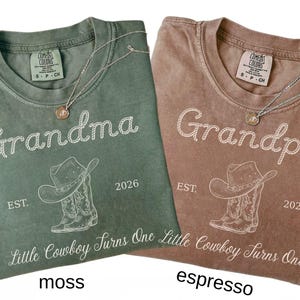 May include: Two short-sleeve t-shirts in moss green and espresso brown. Each shirt features a white graphic of a cowboy hat and boots, the text "Grandma" or "Grandpa," "EST. 2026," and the phrase "Little Cowboy Turns One" in script font.
