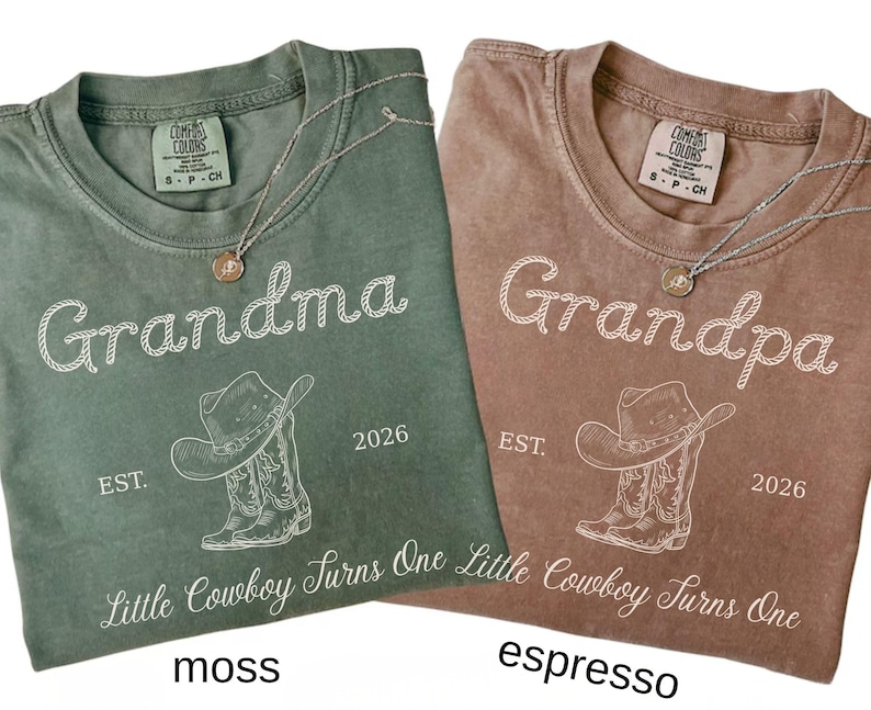 May include: Two short-sleeve t-shirts in moss green and espresso brown. Each shirt features a white graphic of a cowboy hat and boots, the text "Grandma" or "Grandpa," "EST. 2026," and the phrase "Little Cowboy Turns One" in script font.