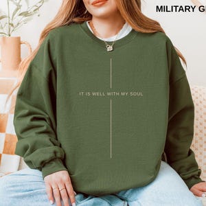 May include: Military green sweatshirt with the text "IT IS WELL WITH MY SOUL" in white. The sweatshirt has a simple, minimalist design with a vertical line graphic. The model is wearing a white undershirt and gold necklace.