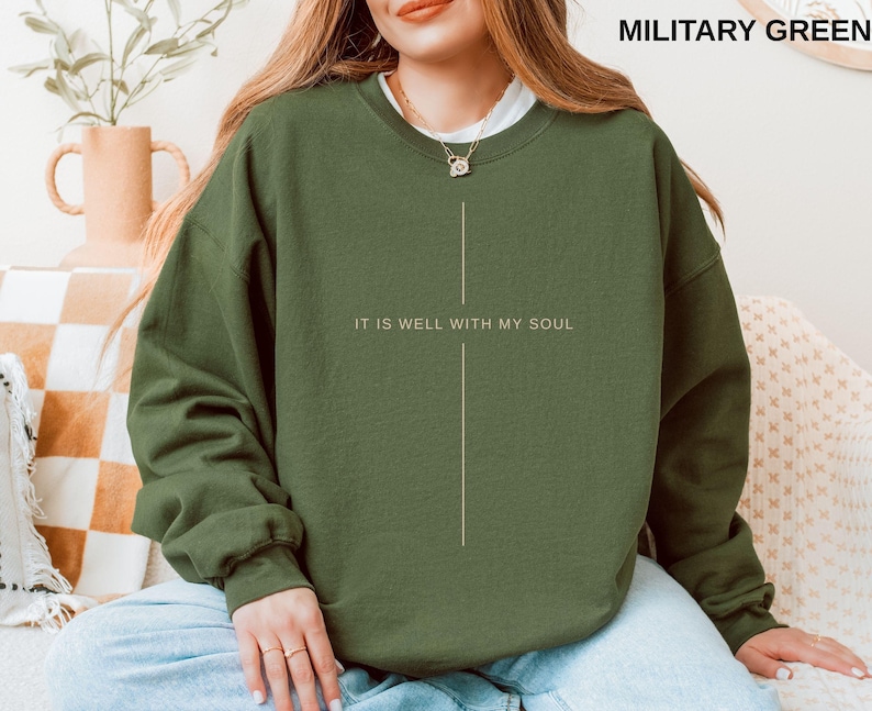 May include: Military green sweatshirt with the text "IT IS WELL WITH MY SOUL" in white. The sweatshirt has a simple, minimalist design with a vertical line graphic. The model is wearing a white undershirt and gold necklace.
