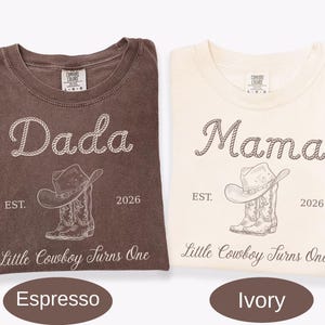 May include: Two t-shirts in espresso and ivory colors. The espresso shirt features white text reading Dada, EST. 2026, and Little Cowboy Turns One, with a cowboy hat and boots graphic. The ivory shirt features brown text and the same graphic.