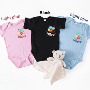 Pu&ograve; includere: Tre body a maniche corte per neonati in rosa chiaro, nero e azzurro. Ognuno presenta una grafica di palloncini colorati con il testo "IT'S FUN TO BE" e il nome "Oliver" stampato sotto. &Egrave; incluso un piccolo orsacchiotto di peluche beige.