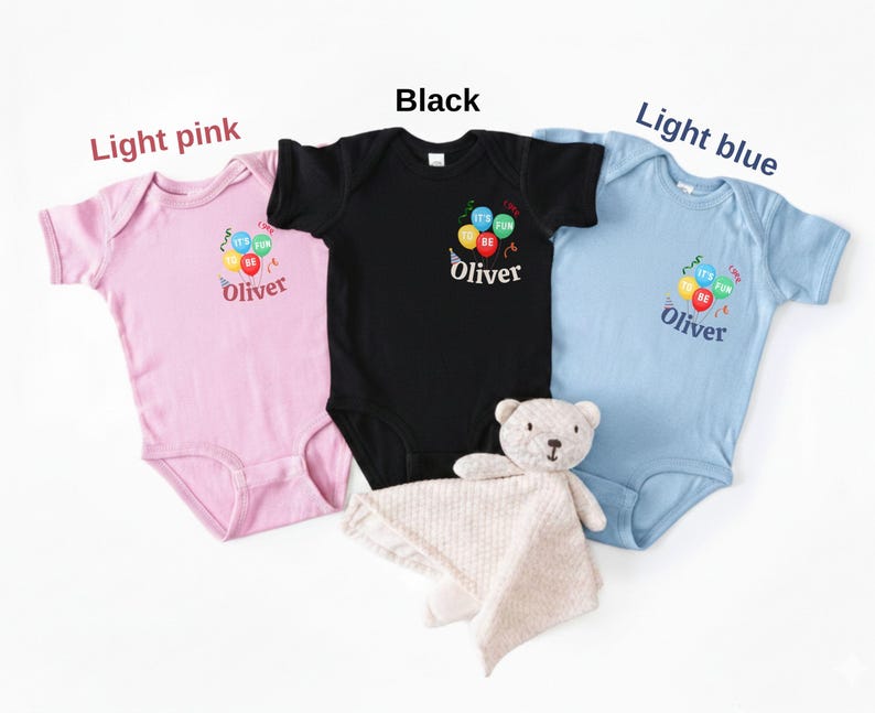 Pu&ograve; includere: Tre body a maniche corte per neonati in rosa chiaro, nero e azzurro. Ognuno presenta una grafica di palloncini colorati con il testo "IT'S FUN TO BE" e il nome "Oliver" stampato sotto. &Egrave; incluso un piccolo orsacchiotto di peluche beige.