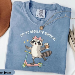 May include: Blue jean colored t-shirt with the text "OFF TO REGULATE EMOTIONS". The shirt features a cartoon raccoon with a pink bow, holding a coffee cup, and riding a pink scooter. The shirt has a Comfort Colors tag.