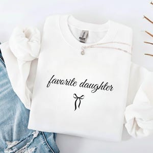 Puede incluir: Una sudadera blanca con la frase "favorite daughter" en fuente negra, con un lazo debajo. Un collar plateado con un peque&ntilde;o colgante descansa sobre la camisa. La sudadera est&aacute; junto a unos vaqueros azules.