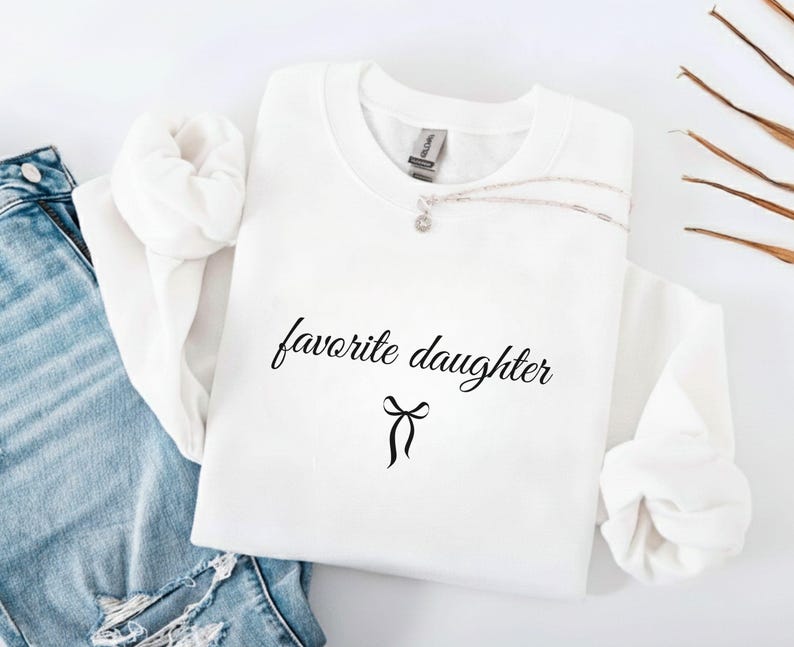 Puede incluir: Una sudadera blanca con la frase "favorite daughter" en fuente negra, con un lazo debajo. Un collar plateado con un peque&ntilde;o colgante descansa sobre la camisa. La sudadera est&aacute; junto a unos vaqueros azules.