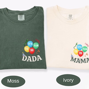 Pu&ograve; includere: Una t-shirt verde muschio e una t-shirt color avorio, entrambe con una grafica di palloncini e stelle filanti colorate. La maglia verde riporta la scritta "IT'S FUN TO BE DADA" e quella avorio "IT'S FUN TO BE MAMA" in bianco e grigio scuro.