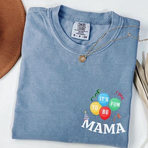 Pu&ograve; includere: Una t-shirt blu a maniche corte con scollo rotondo ed etichetta Comfort Colors. La parte anteriore presenta una grafica di palloncini colorati in giallo, blu, rosso e verde con il testo 'IT'S FUN TO BE MAMA' in lettere bianche sotto i palloncini.