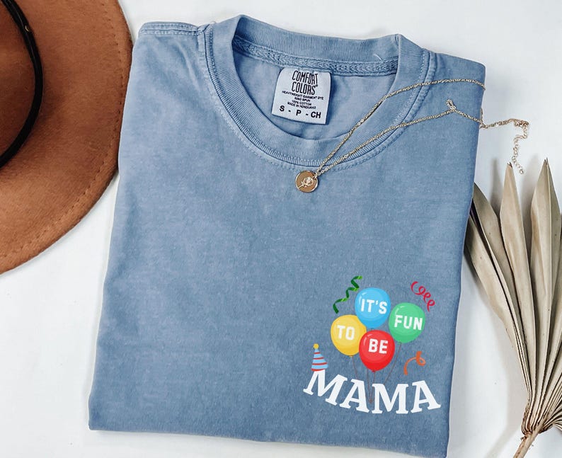 Pu&ograve; includere: Una t-shirt blu a maniche corte con scollo rotondo ed etichetta Comfort Colors. La parte anteriore presenta una grafica di palloncini colorati in giallo, blu, rosso e verde con il testo 'IT'S FUN TO BE MAMA' in lettere bianche sotto i palloncini.