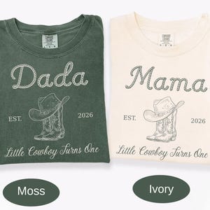 May include: Two short-sleeve crew neck t-shirts in moss green and ivory. The moss shirt features white text reading Dada, and the ivory shirt features dark text reading Mama. Both include a cowboy hat, boots, EST. 2026, and Little Cowboy Turns One.