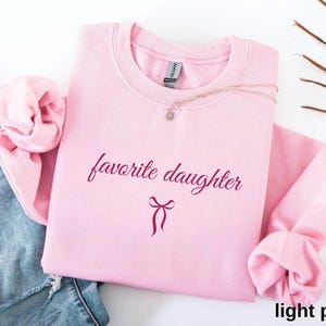 Puede incluir: Una sudadera rosa claro con la frase "favorite daughter" en una fuente burdeos. Un gr&aacute;fico de lazo burdeos est&aacute; debajo del texto. La sudadera est&aacute; doblada, con las mangas y el cuello visibles. Un collar con un peque&ntilde;o colgante est&aacute; sobre la sudadera.