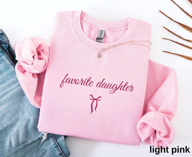 Puede incluir: Una sudadera rosa claro con la frase "favorite daughter" en una fuente burdeos. Un gr&aacute;fico de lazo burdeos est&aacute; debajo del texto. La sudadera est&aacute; doblada, con las mangas y el cuello visibles. Un collar con un peque&ntilde;o colgante est&aacute; sobre la sudadera.
