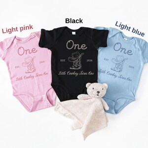 May include: Three short-sleeve baby bodysuits in light pink, black, and light blue. Each features a graphic of a cowboy hat and boots with the text One, EST. 2026, and Little Cowboy Turns One. A small, light-colored plush teddy bear is included.
