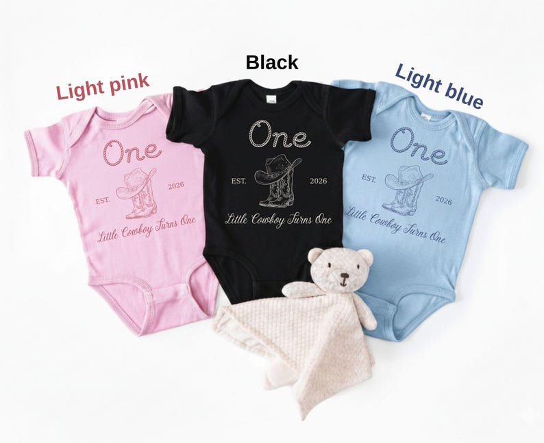 May include: Three short-sleeve baby bodysuits in light pink, black, and light blue. Each features a graphic of a cowboy hat and boots with the text One, EST. 2026, and Little Cowboy Turns One. A small, light-colored plush teddy bear is included.