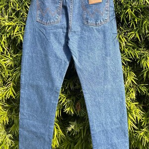 May include: Blue denim jeans with classic five-pocket styling. Features include a brown leather patch with the brand name, orange stitching, and the iconic 'W' stitching on the back pockets. The jeans are displayed against a backdrop of green foliage.