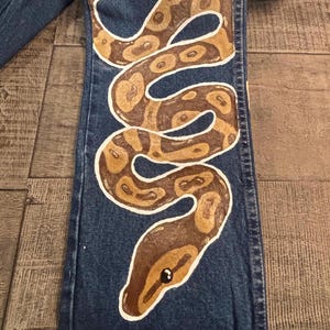 May include: Blue denim jeans with a hand-painted snake design. The snake is brown and tan with white outlines, coiled down the leg of the jeans. The snake's head is at the bottom, with a black eye.