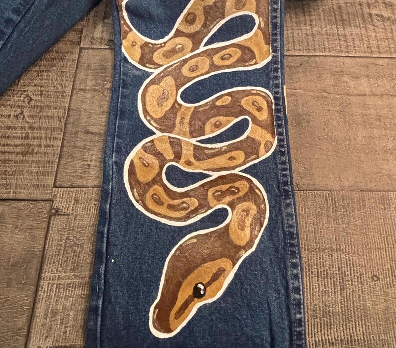 May include: Blue denim jeans with a hand-painted snake design. The snake is brown and tan with white outlines, coiled down the leg of the jeans. The snake's head is at the bottom, with a black eye.