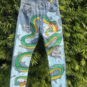 Puede incluir: Vaqueros de mezclilla azul claro con un diseño de dragón verde y amarillo pintado a mano. El dragón serpentea alrededor de las piernas, sobre un fondo de nubes azules. Los vaqueros tienen detalles desgastados.