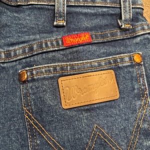 May include: Close-up of blue denim jeans with the Wrangler logo on the back pocket and a red Wrangler tag. The jeans have classic stitching and copper-colored rivets. The overall look is vintage and casual.