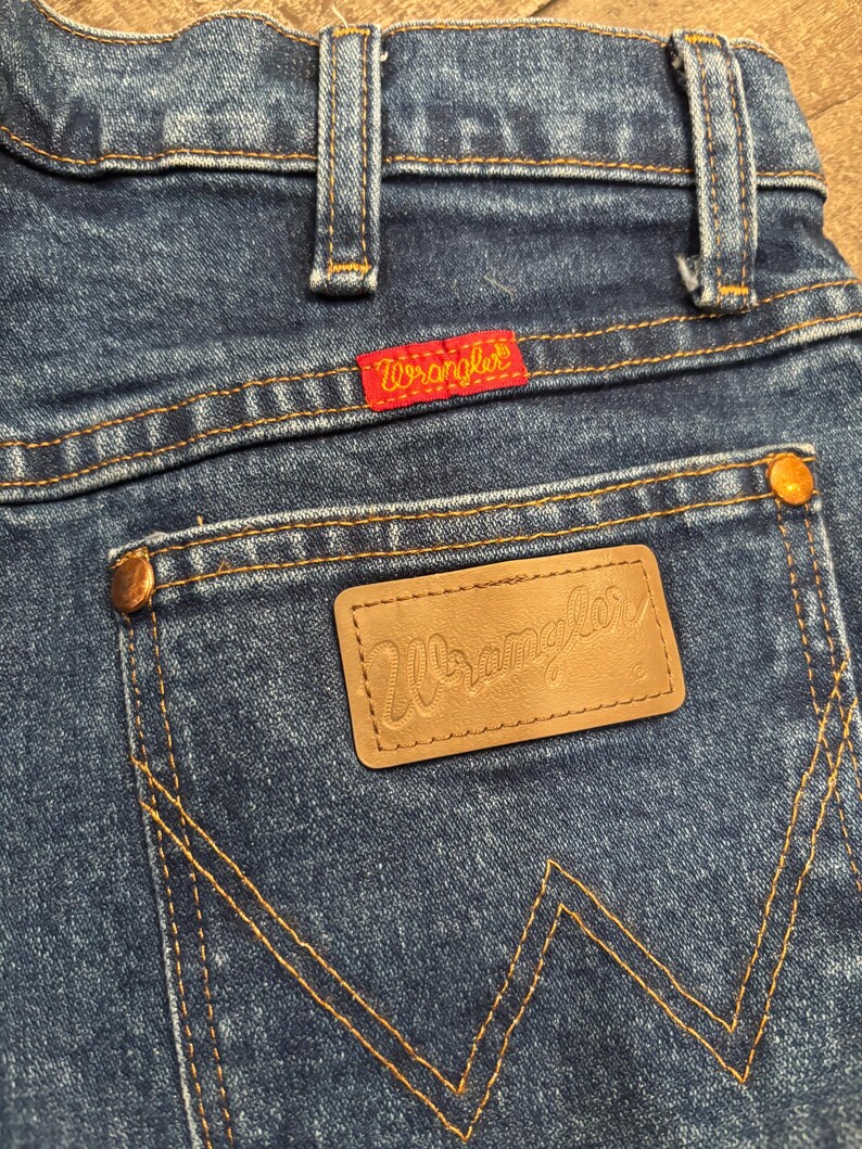 May include: Close-up of blue denim jeans with the Wrangler logo on the back pocket and a red Wrangler tag. The jeans have classic stitching and copper-colored rivets. The overall look is vintage and casual.