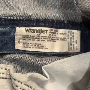 May include: Close-up of a Wrangler jeans label. The label is white with black text, including washing instructions and size 11 x 34. The jeans are blue denim, with visible stitching and the inside of the pocket.
