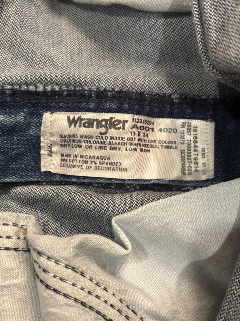 May include: Close-up of a Wrangler jeans label. The label is white with black text, including washing instructions and size 11 x 34. The jeans are blue denim, with visible stitching and the inside of the pocket.
