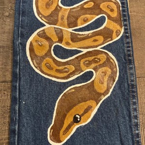 May include: Blue denim jeans with a hand-painted snake design. The snake is brown and tan with white outlines, coiled across the leg. The snake's head is at the bottom, with a black eye.