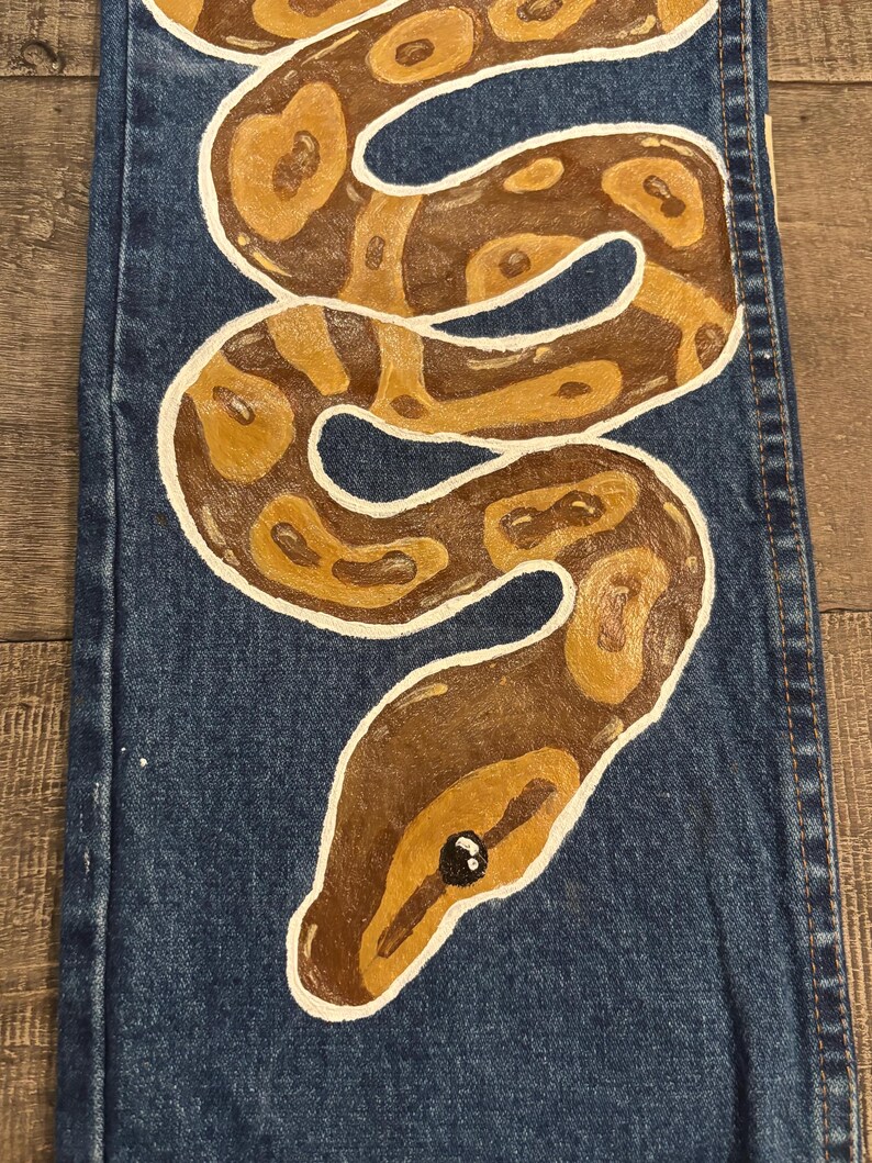 May include: Blue denim jeans with a hand-painted snake design. The snake is brown and tan with white outlines, coiled across the leg. The snake's head is at the bottom, with a black eye.