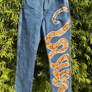 May include: Blue denim jeans with a hand-painted snake design in shades of brown and cream. The snake winds down the front of the jeans, creating a unique and artistic look. The jeans have a classic five-pocket design.