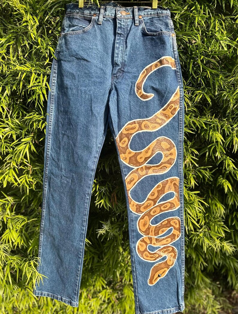 May include: Blue denim jeans with a hand-painted snake design in shades of brown and cream. The snake winds down the front of the jeans, creating a unique and artistic look. The jeans have a classic five-pocket design.