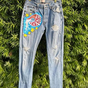 Puede incluir: Vaqueros de mezclilla azul claro con detalles desgastados y un diseño pintado. El diseño presenta una concha de nautilus con rayas rojas y blancas, detalles azules y estrellas amarillas. Los vaqueros cuelgan de una percha blanca.