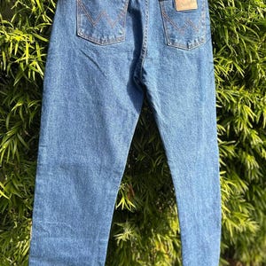 May include: Blue denim jeans with classic five-pocket styling. Features include copper rivets, a leather patch, and the Wrangler logo. The back pockets have the signature 'W' stitching. The jeans are hanging against a backdrop of green foliage.