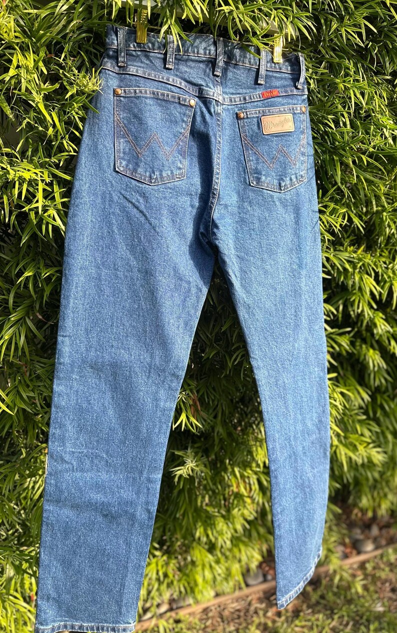 May include: Blue denim jeans with classic five-pocket styling. Features include copper rivets, a leather patch, and the Wrangler logo. The back pockets have the signature 'W' stitching. The jeans are hanging against a backdrop of green foliage.