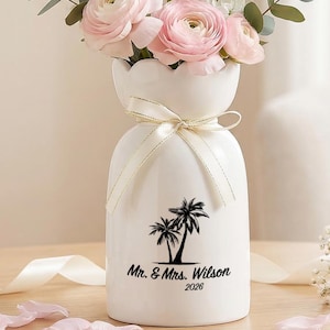 May include: A white ceramic vase with a scalloped rim, tied with a cream ribbon, featuring a black graphic of two palm trees and the text "Mr. & Mrs. Wilson 2026". The vase is filled with a bouquet of light pink ranunculus and white flowers.