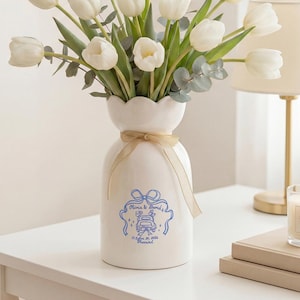 以下が含まれることがあります： 白い波型の縁が付いた白いセラミック花瓶には、白いチューリップと緑のユーカリの葉のブーケが入っています。 花瓶は青と白のデザインで、ベージュのリボンで結ばれています。 火の灯ったアロマキャンドルとランプが背景にあります。